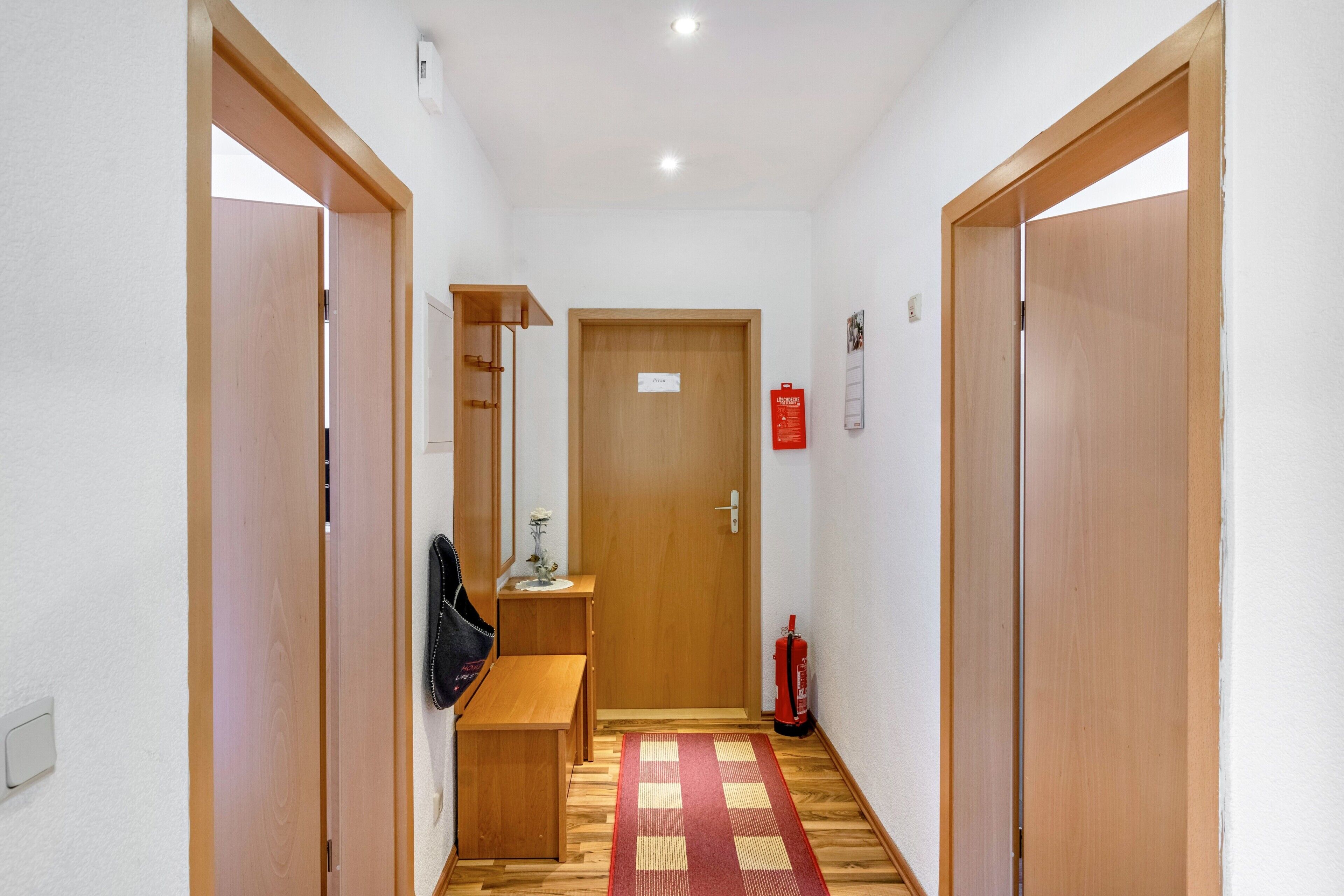A hallway with wooden doors, a bench, a small table with a vase, a fire extinguisher, and a sign on the door.