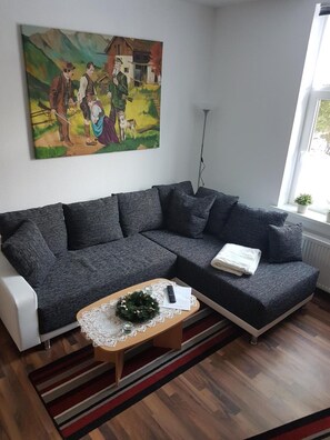 Ett modernt vardagsrum med en grå hörnsoffa, ett träbord och en spetsduk på bordet, samt en tavla med ett lantligt motiv på väggen.