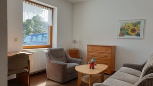 Ein Zimmer mit Fenster, Heizkörper, Lampe, Kommode, Stuhl, Sofa und kleinem Tisch.