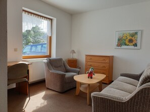 A room with a window, a radiator, a lamp, a dresser, a chair, a sofa, and a small table. - "Wiesengrund" Apartment, located on the ground floor of the Werraquelle building, which is ideal for groups (Neuhaus am Rennweg)