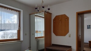 Ein Schlafzimmer mit einem Bett, einem Fenster mit Vorhängen, einer hölzernen Bank, einem Spiegel und einer Tür zu einem weiteren Raum.