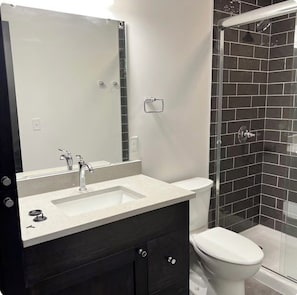 A bathroom with a mirror, sink, dark wooden vanity, white toilet, and a shower area with black tiles. - Brick & Bed - 2 Bedroom Riverfront Loft (St. Louis)
