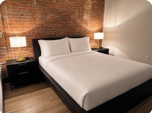 A bed with white linens, two bedside tables with lamps, a brick wall, and a wooden floor. - Brick & Bed - 2 Bedroom Riverfront Loft (St. Louis)