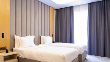 Een hotelkamer met twee bedden, een houten nachtkastje, een lade en een groot raam met gordijnen.