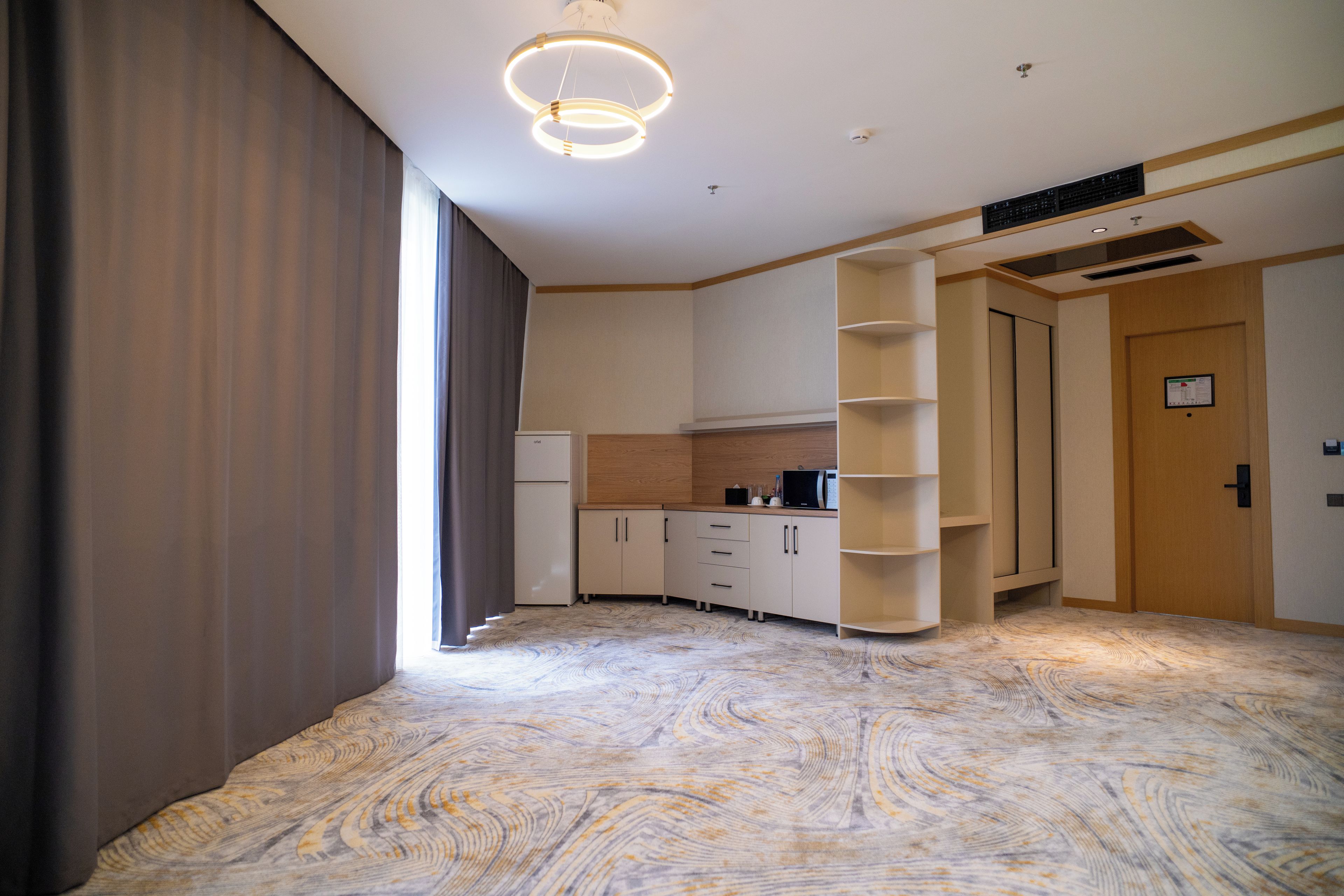 A hotel room with a modern design, featuring a built-in kitchen, a wardrobe, and a large window with curtains.