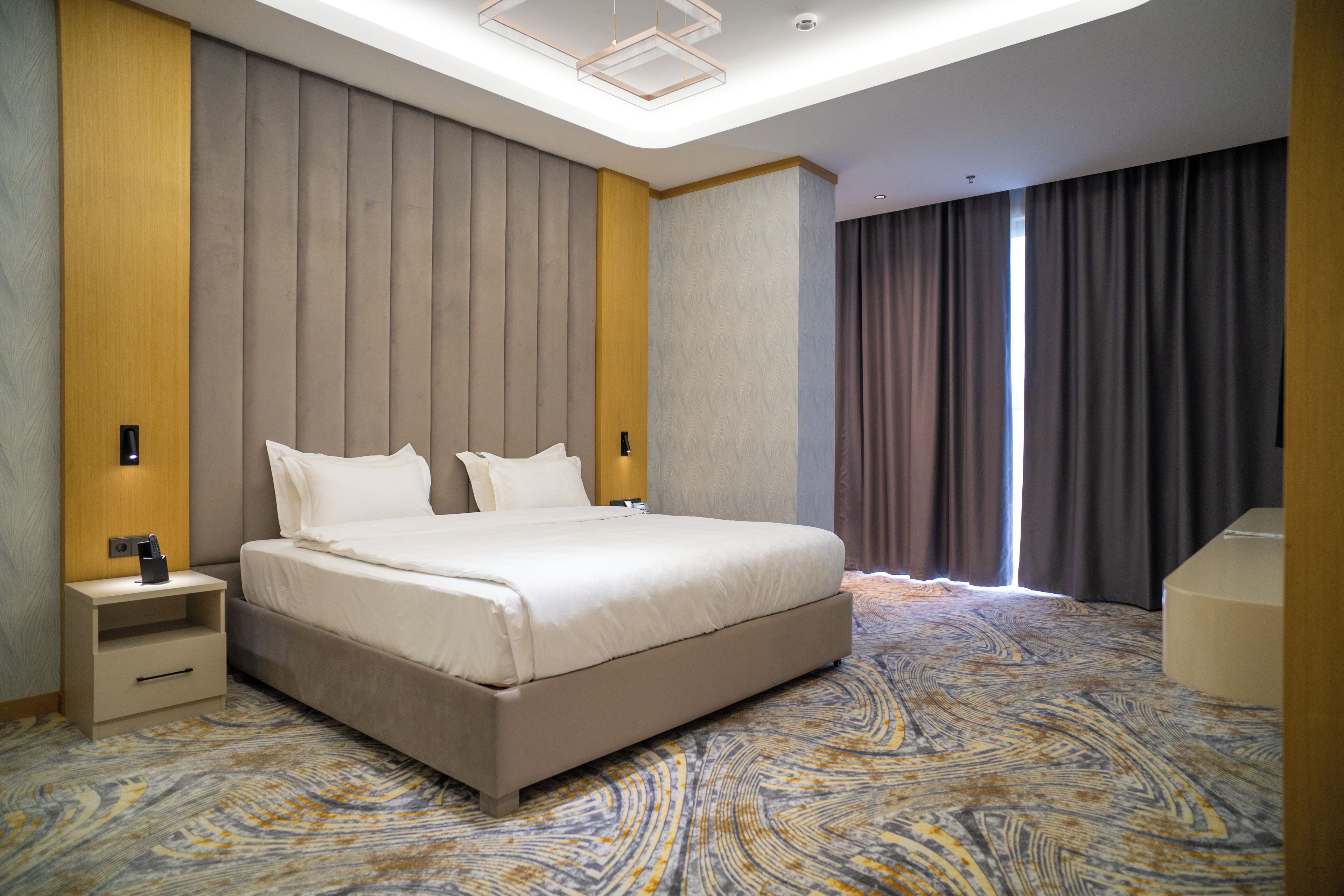 Una habitación de hotel moderna con una cama grande, una mesita de noche, un escritorio integrado y un amplio ventanal con cortinas.
