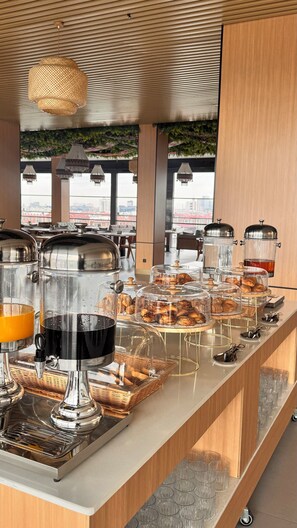 Um buffet de café da manhã com diversos alimentos e bebidas, teto de madeira e vista para edifícios ao fundo.