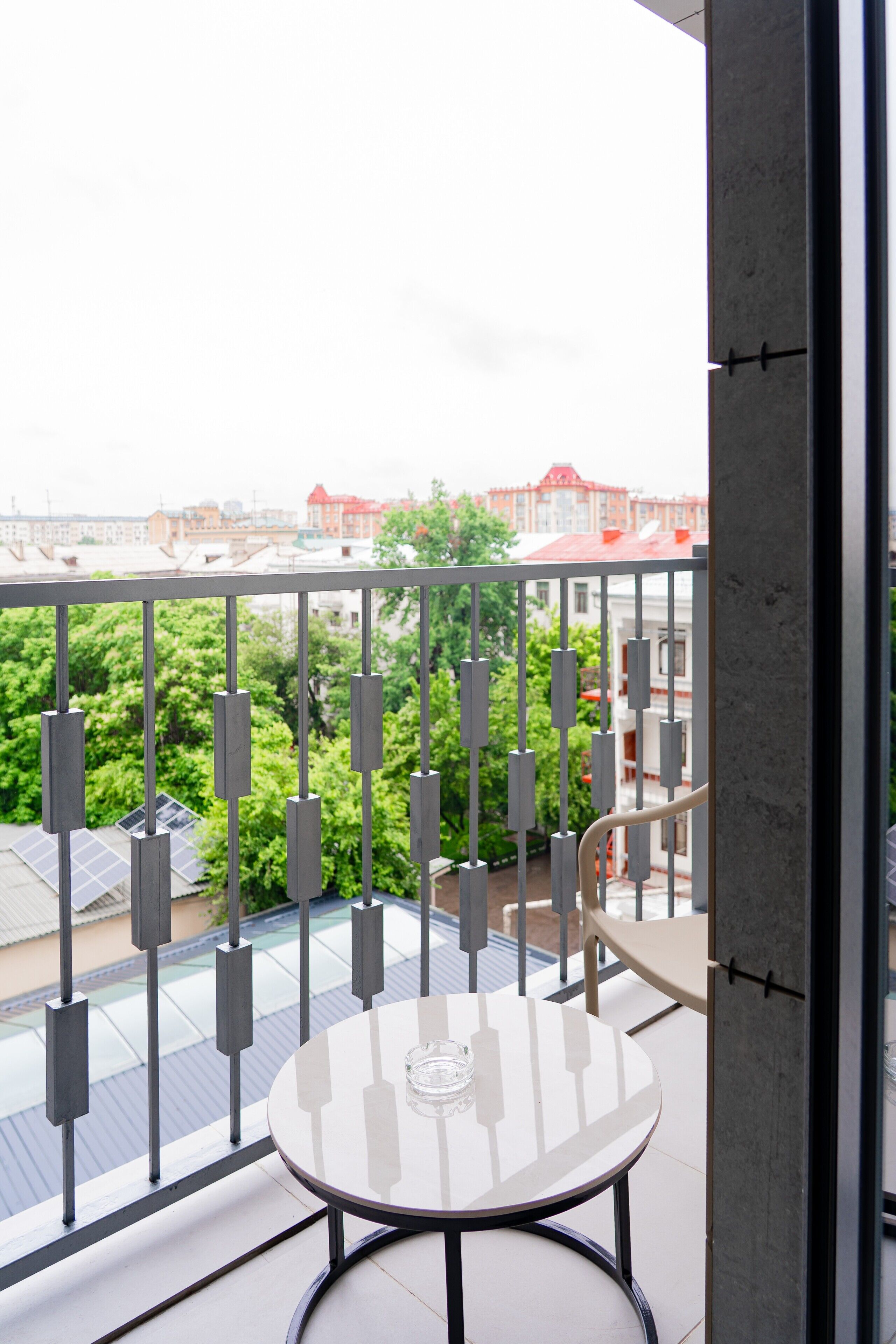 En balkon med et bord og stole, et gelænder i metal og udsigt til bygninger og træer.