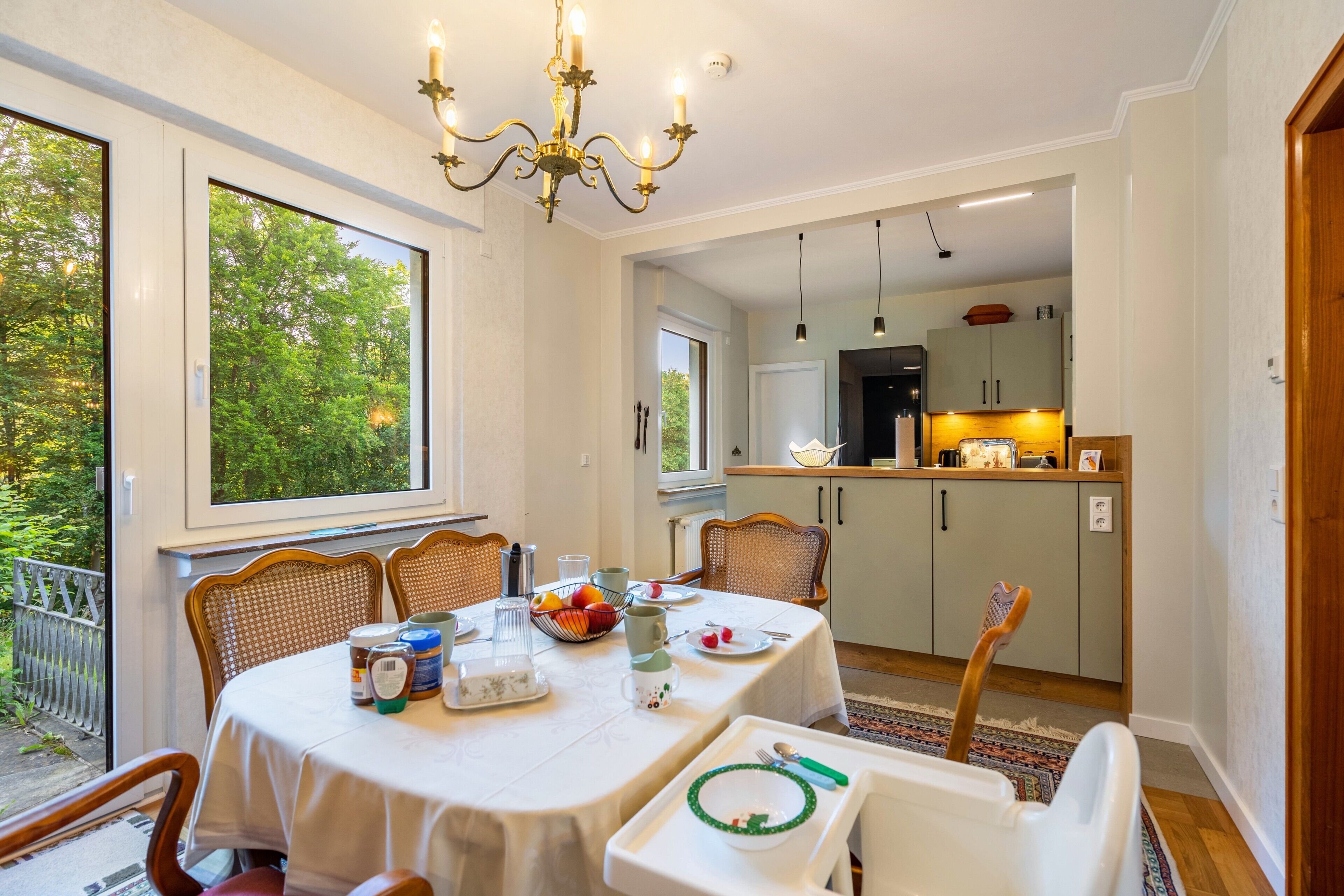 A dining area with a table set for four, chairs, a chandelier, and a view of the kitchen in the background.