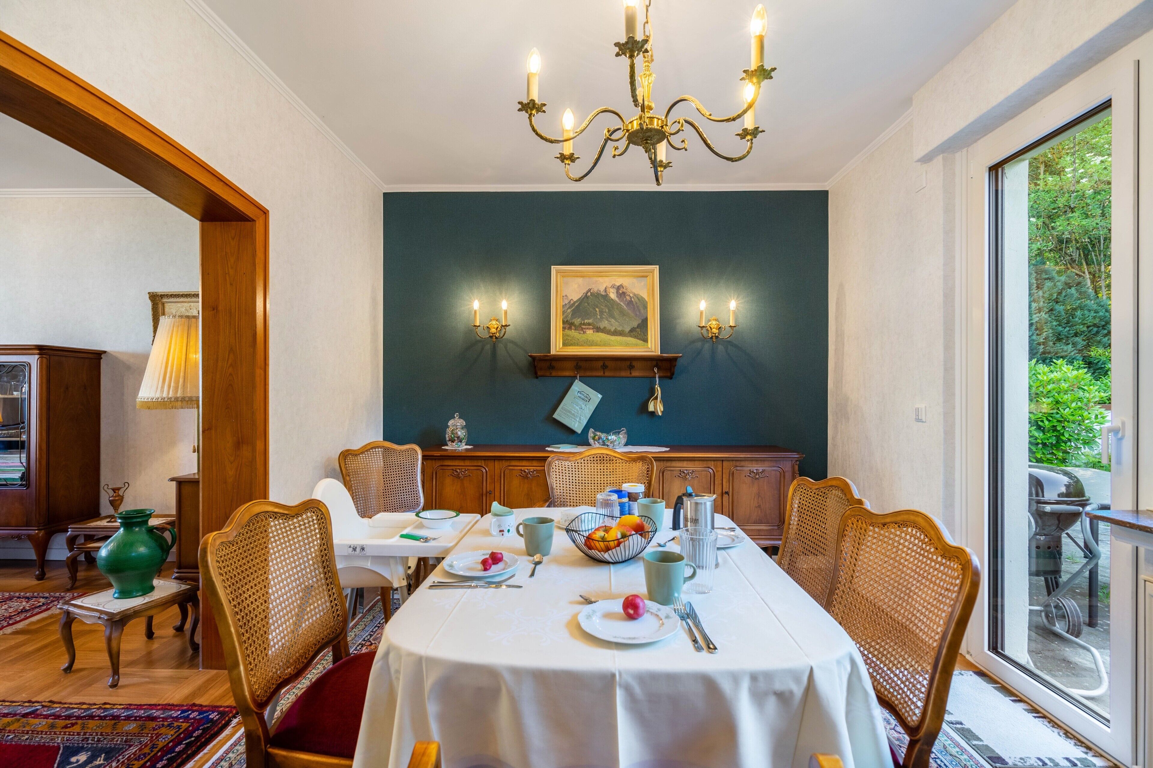 A dining room with a table set for four, a chandelier, and a view through a window.
