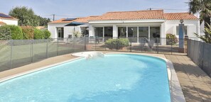 Ein Schwimmbad mit Glaszaun, ein weißes Gebäude mit einem Ziegeldach und ein klarer Himmel.