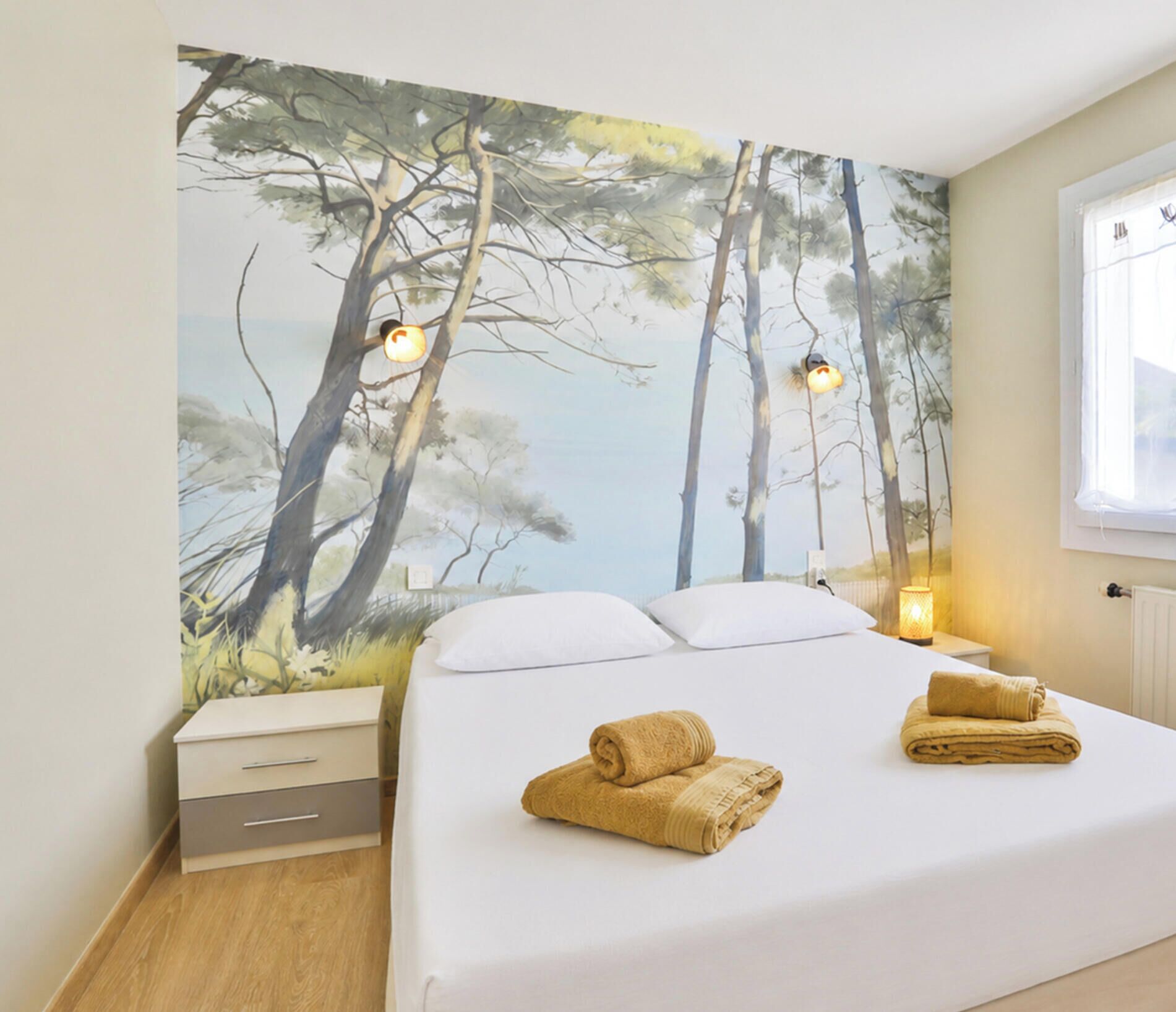 Une chambre à coucher avec un lit, deux tables de chevet, une petite table et une fresque murale représentant des arbres.
