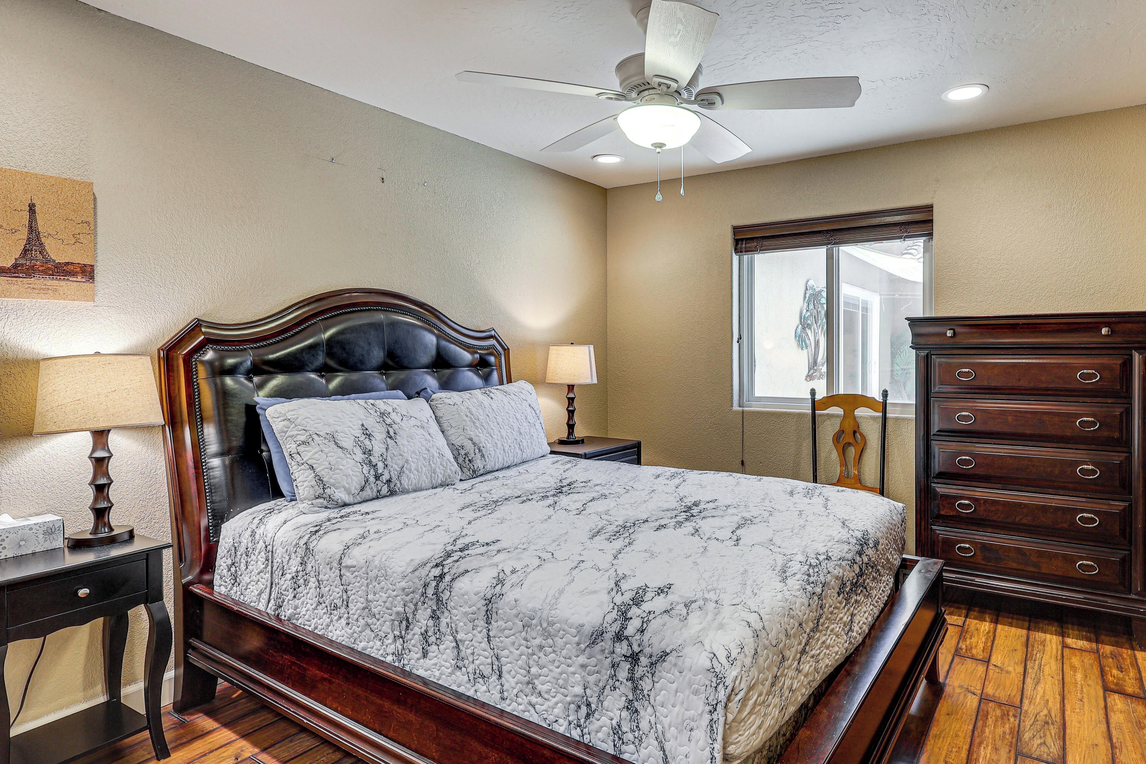 A bedroom with a bed, a dresser, a nightstand, a lamp, a ceiling fan, and a window.