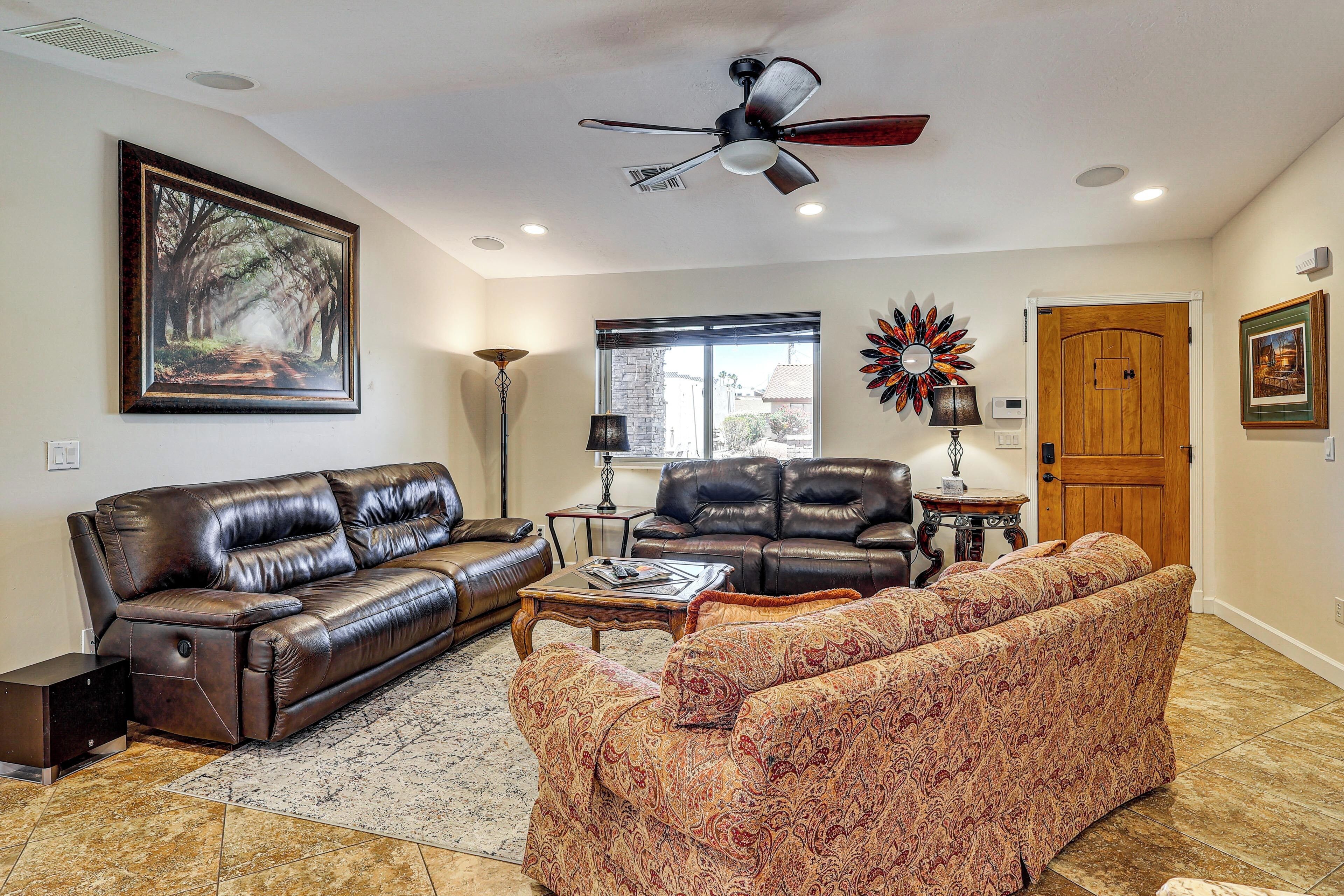A living room with a brown leather sofa, a patterned armchair, a ceiling fan, and a large window.