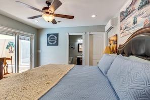 A bedroom with a bed, ceiling fan, artwork, and a view of the outside.