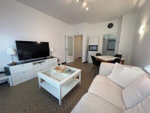 Ein modernes Wohnzimmer mit einem Flachbildfernseher, einem Glastisch, einem weißen Sofa, einem Esstisch mit Stühlen und einer Küchenzeile im Hintergrund.