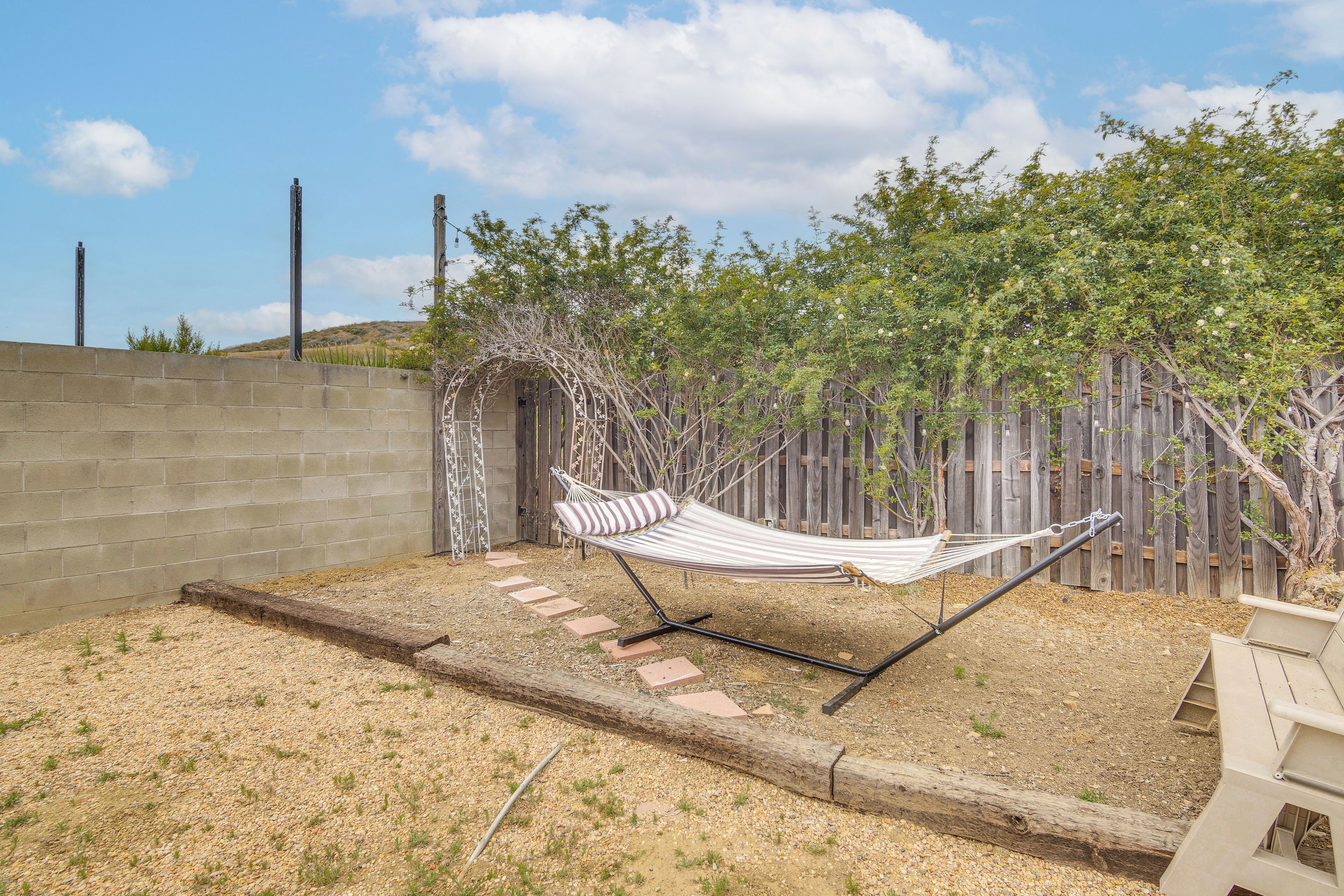 Un hamac dans un jardin, avec une clôture en bois, un sol recouvert de gravier et un chemin pavé de briques.
