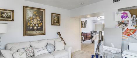 Een woonkamer met een witte bank, een bloemistisch kunstwerk in een lijst, een gitaar en een televisie.