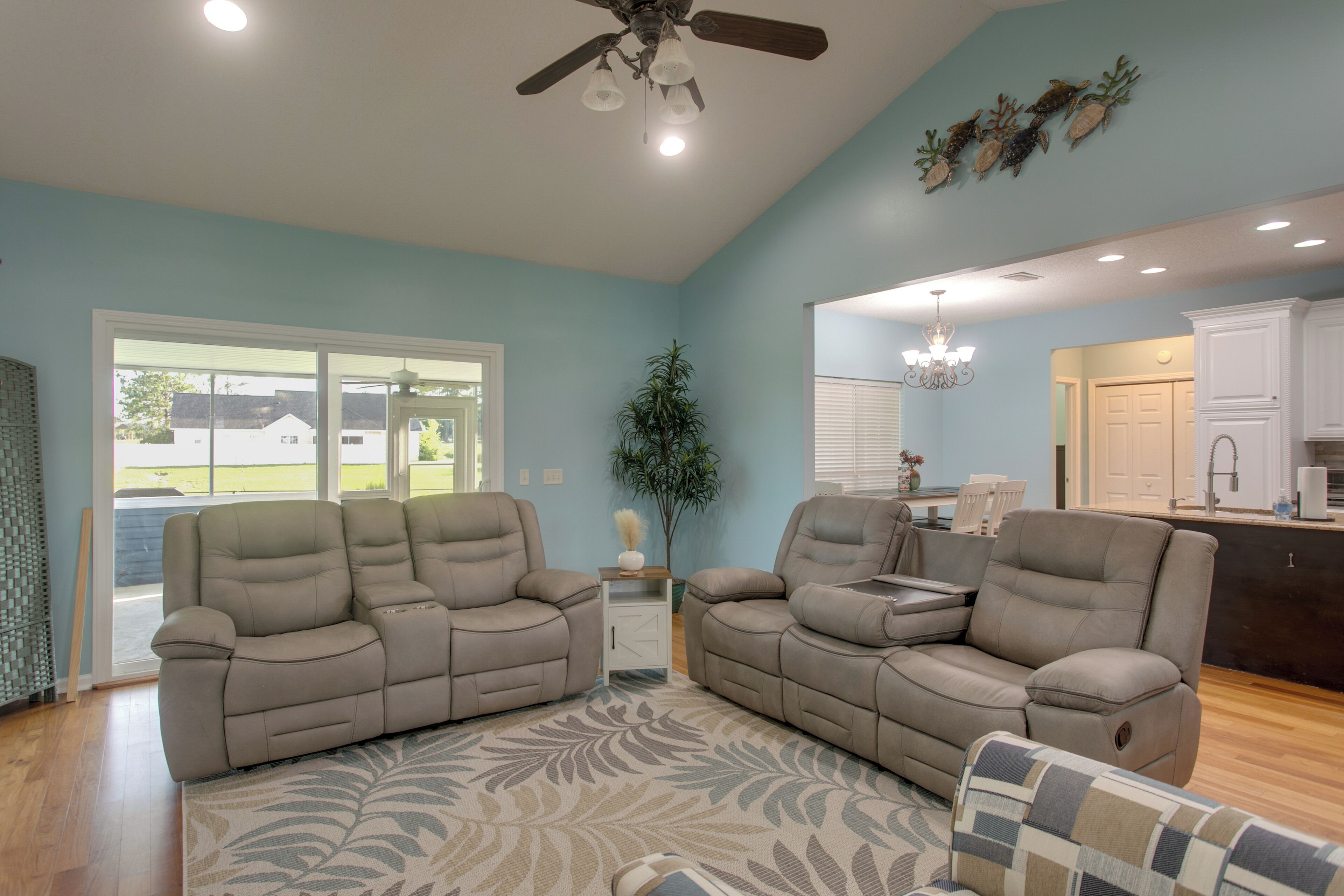 A living room with a large sectional sofa, a small side table, a potted plant, and a view of the kitchen through an open door.