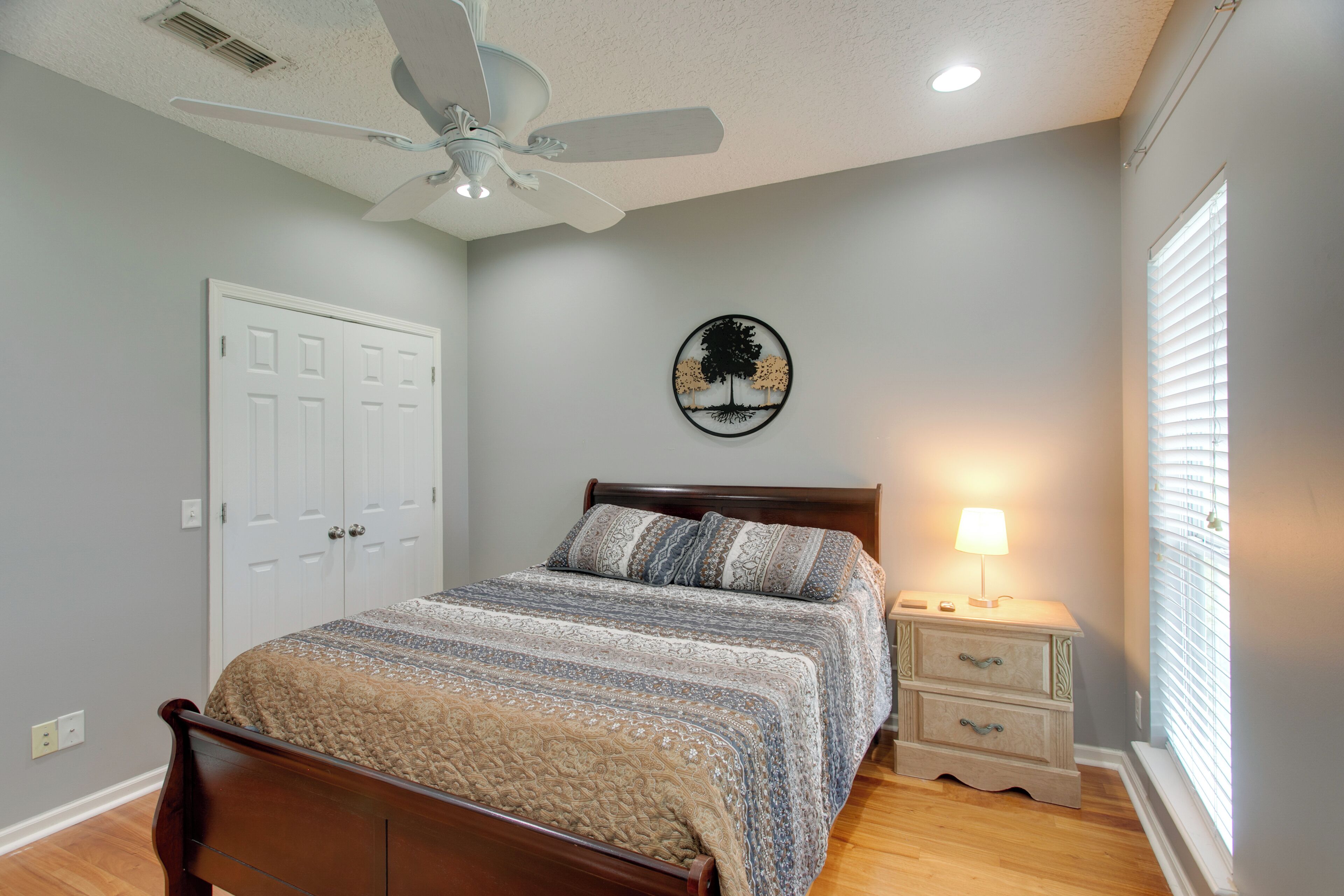A bedroom with a bed, a nightstand, a wall art piece, a ceiling fan, and a window with blinds.