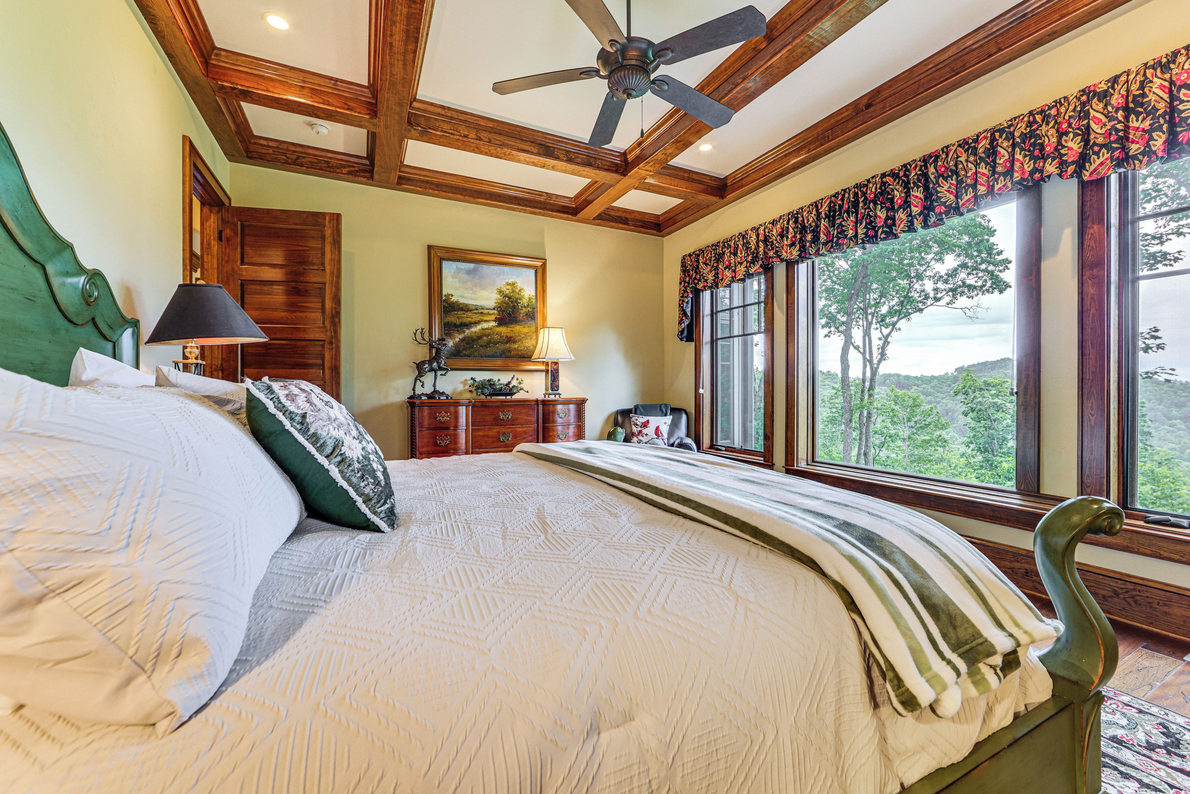 A bedroom with a bed, a ceiling fan, a dresser, a lamp, a painting, and a window with a view.