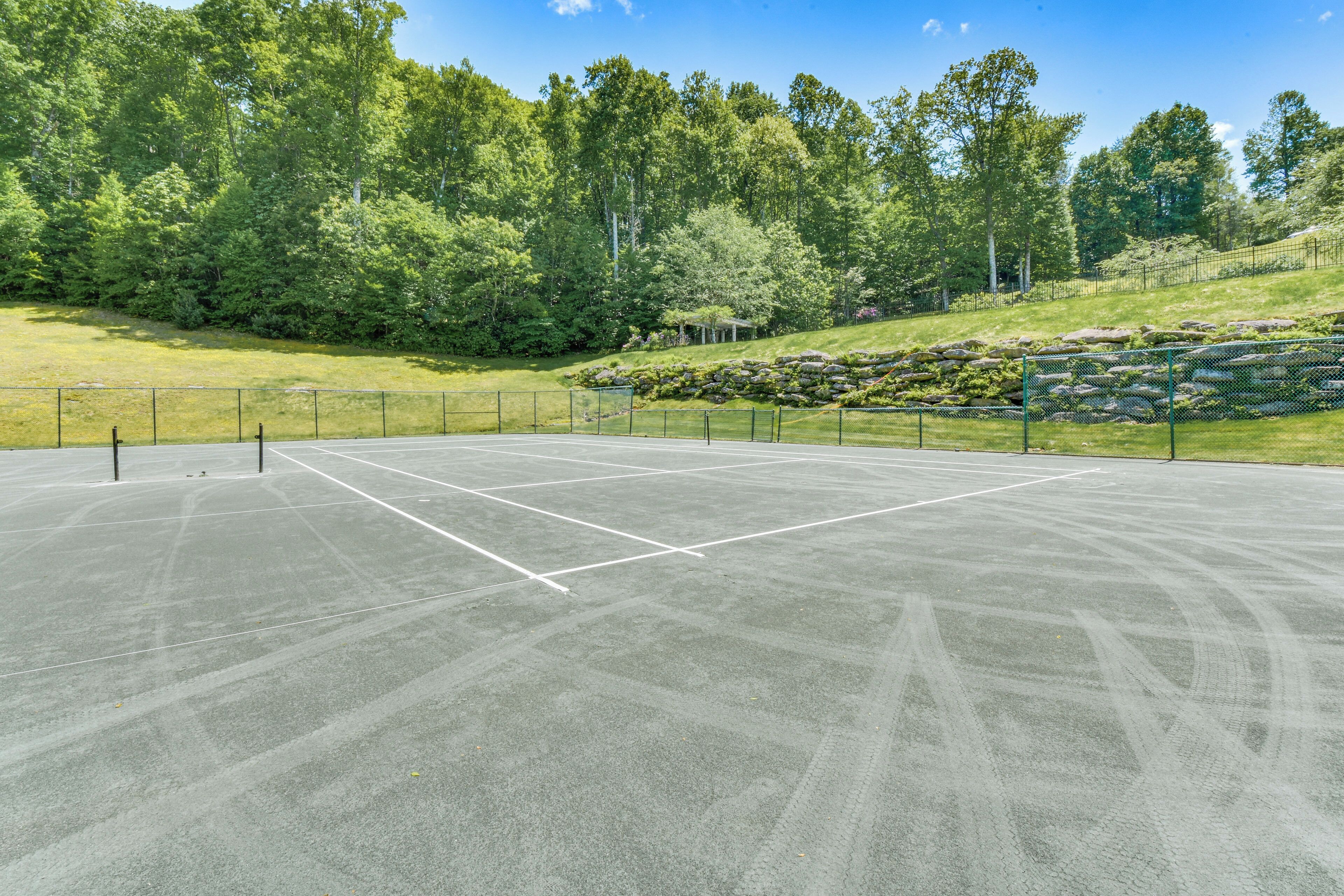 An outdoor tennis court with a net, surrounded by a fence, and a grassy area with trees and rocks in the background.