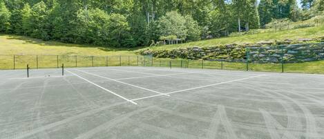 An outdoor tennis court with a net, surrounded by a fence, and a grassy area with trees and rocks in the background.