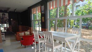 A dining area with white chairs and a table, large windows with striped awnings, and a red chair in the background. - Central Perk Patong (Patong)
