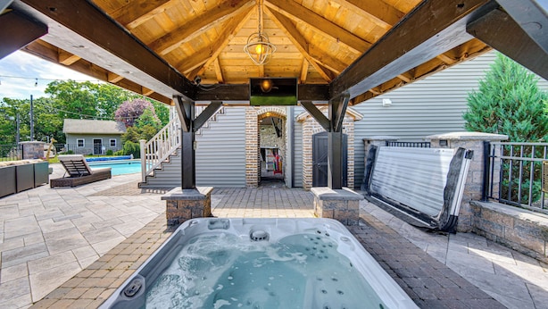 A covered outdoor hot tub area with a wooden ceiling, stone pillars, and a view of a house in the background. - Pool, Hot Tub + Views: Spacious Danville Escape! (Danville)