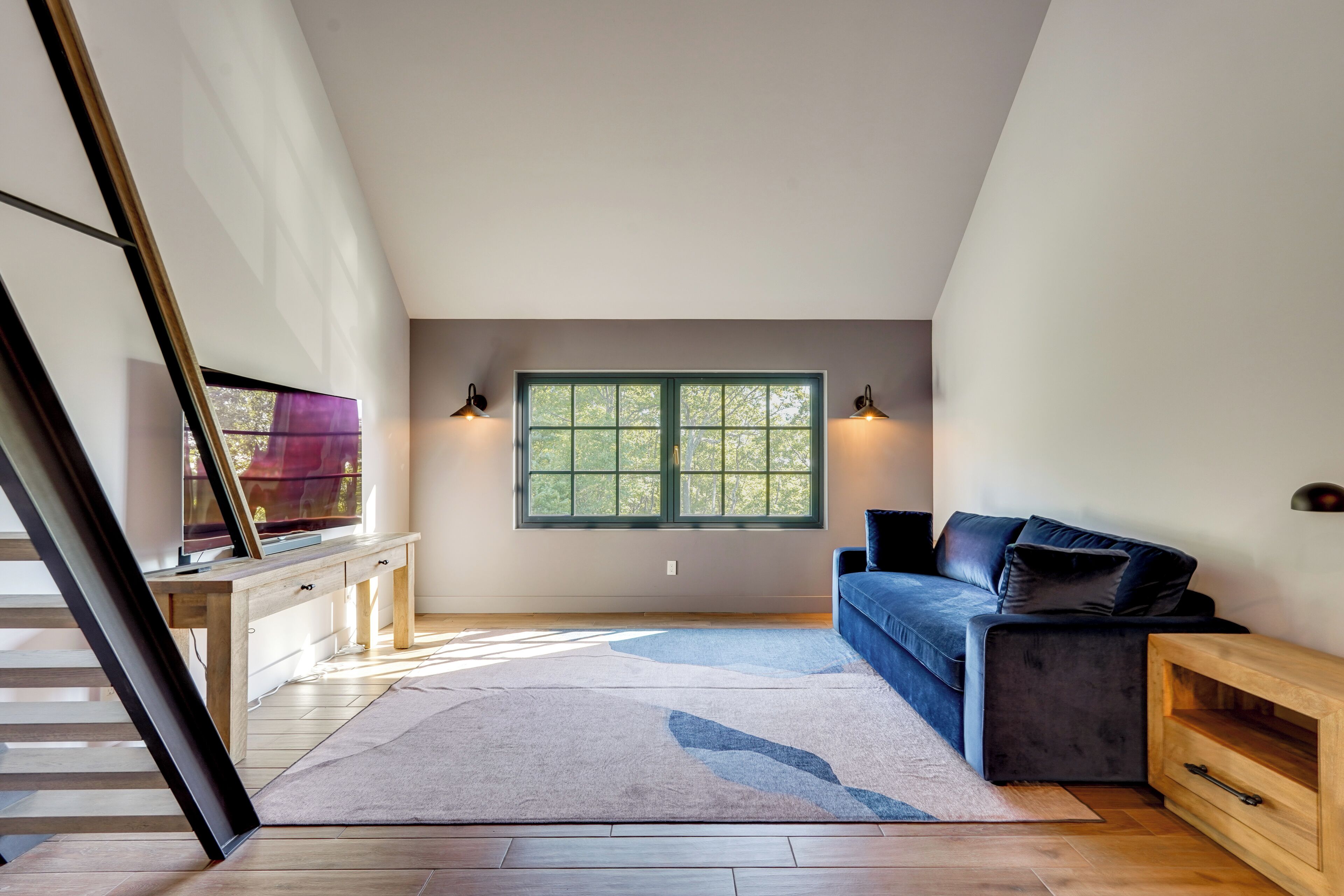 Ein modernes Schlafzimmer im Loft-Stil mit großem Fenster, einem Holztisch, einem dunklen Sofa und einer Treppe.
