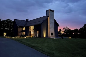 A modern house with a large chimney, large windows, and a well-lit exterior at dusk.