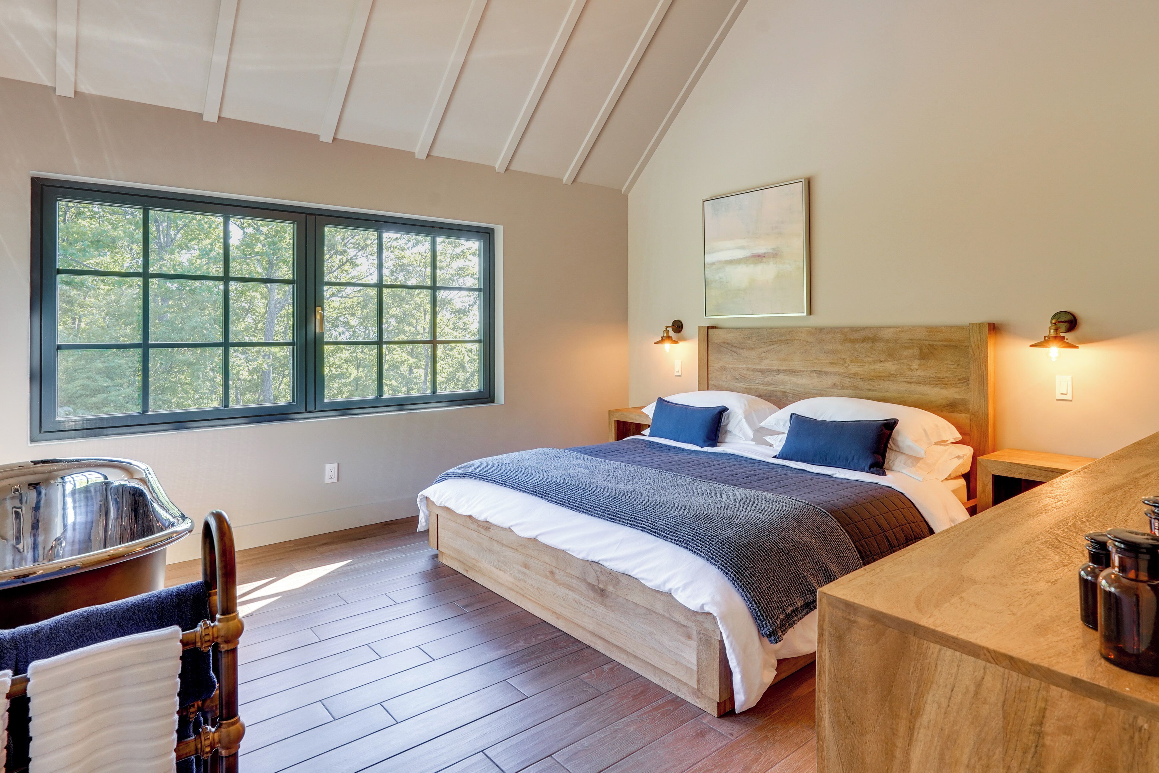 A bedroom with a large window, a wooden bed, a nightstand, a headboard, and a wooden desk with a chair.