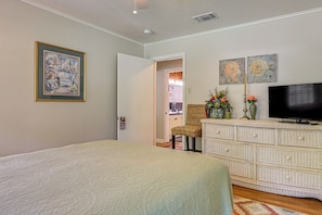 Un dormitorio con cama, televisor, cómoda de mimbre y una puerta que da a otra habitación.