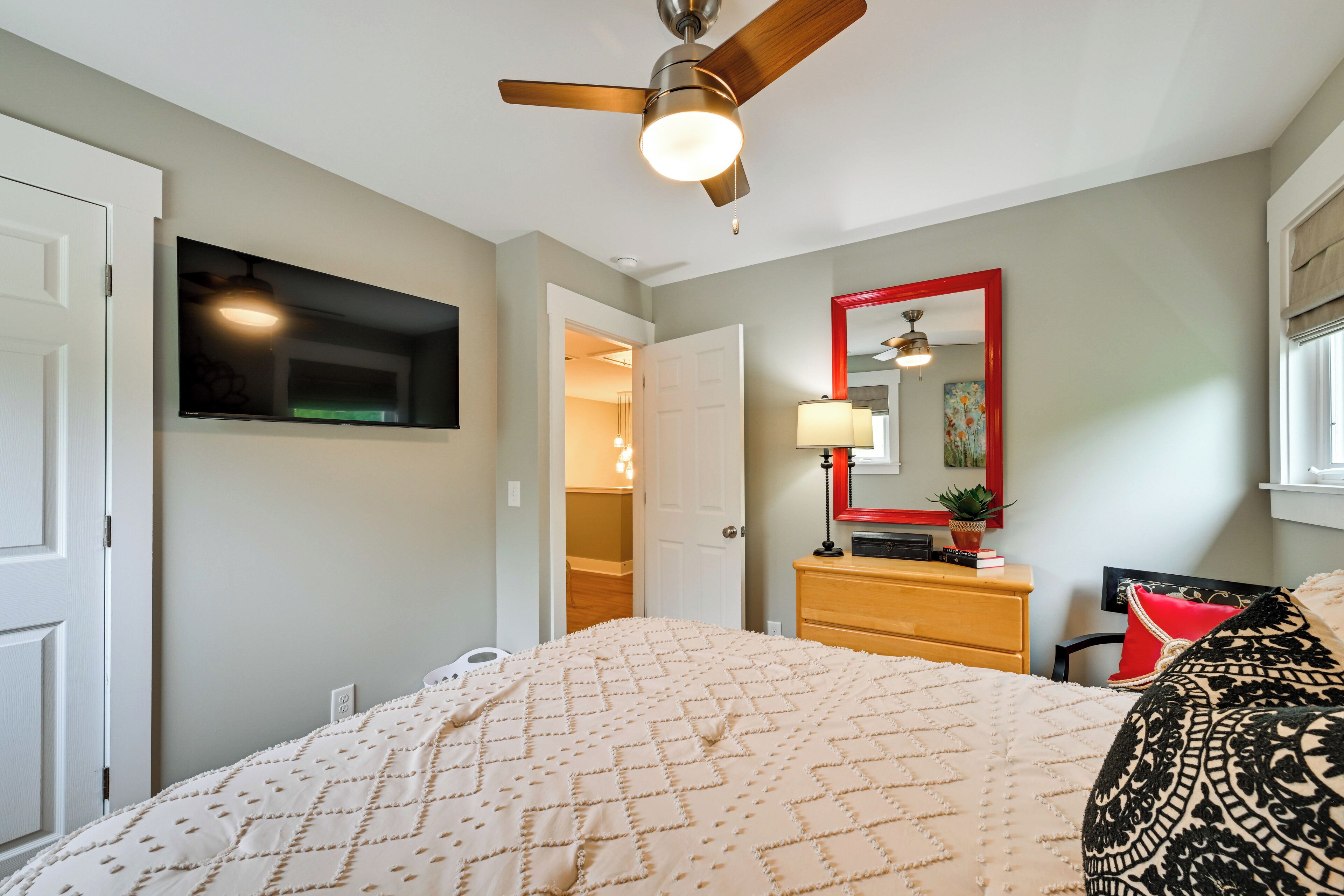 Une chambre à coucher comprenant un lit, un ventilateur de plafond, une commode, un miroir et une porte donnant sur une autre pièce.