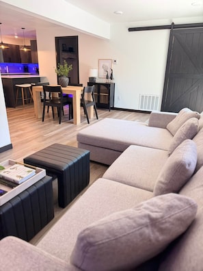 Ein modernes Wohnzimmer mit einer Eckcouch, einem Essbereich mit Stühlen und einem Tisch sowie einer Küche im Hintergrund.