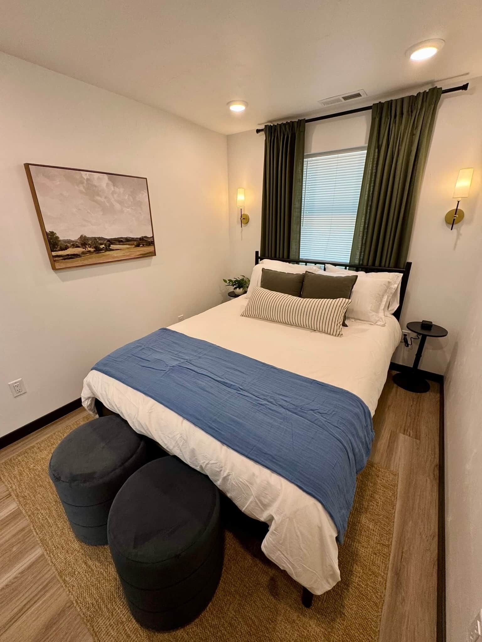 Een keurig opgemaakt slaapkamer met een bed, twee poufs, een nachtkastje, een lamp en een schilderij aan de muur.