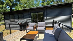 Un patio exterior moderno con terraza de madera, un sofá negro, una mesa de madera y una zona de barbacoa.