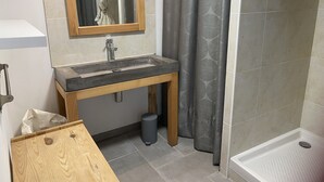 A bathroom with a wooden vanity, a sink, a mirror, a shower with a patterned curtain, and a tiled floor. - Self-contained apartment in the heart of the garrigues. Spa garden. pets allowed (Pignan)