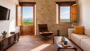 Un soggiorno con parete in pietra, due finestre, una poltrona in pelle marrone, un divano, un tavolino da caffè e una televisione.