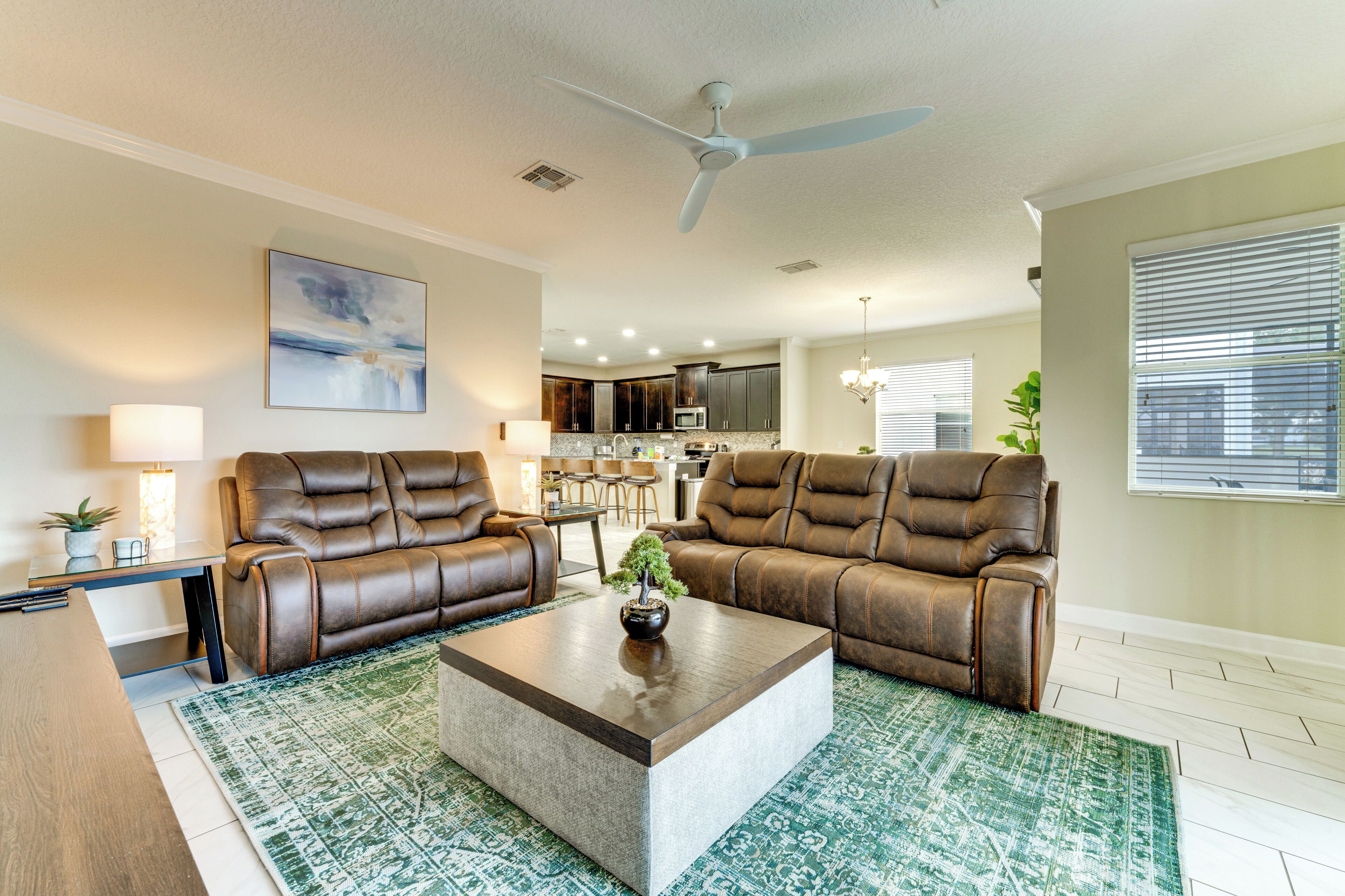 A living room with two brown leather sofas, a coffee table, a ceiling fan, and a kitchen in the background.