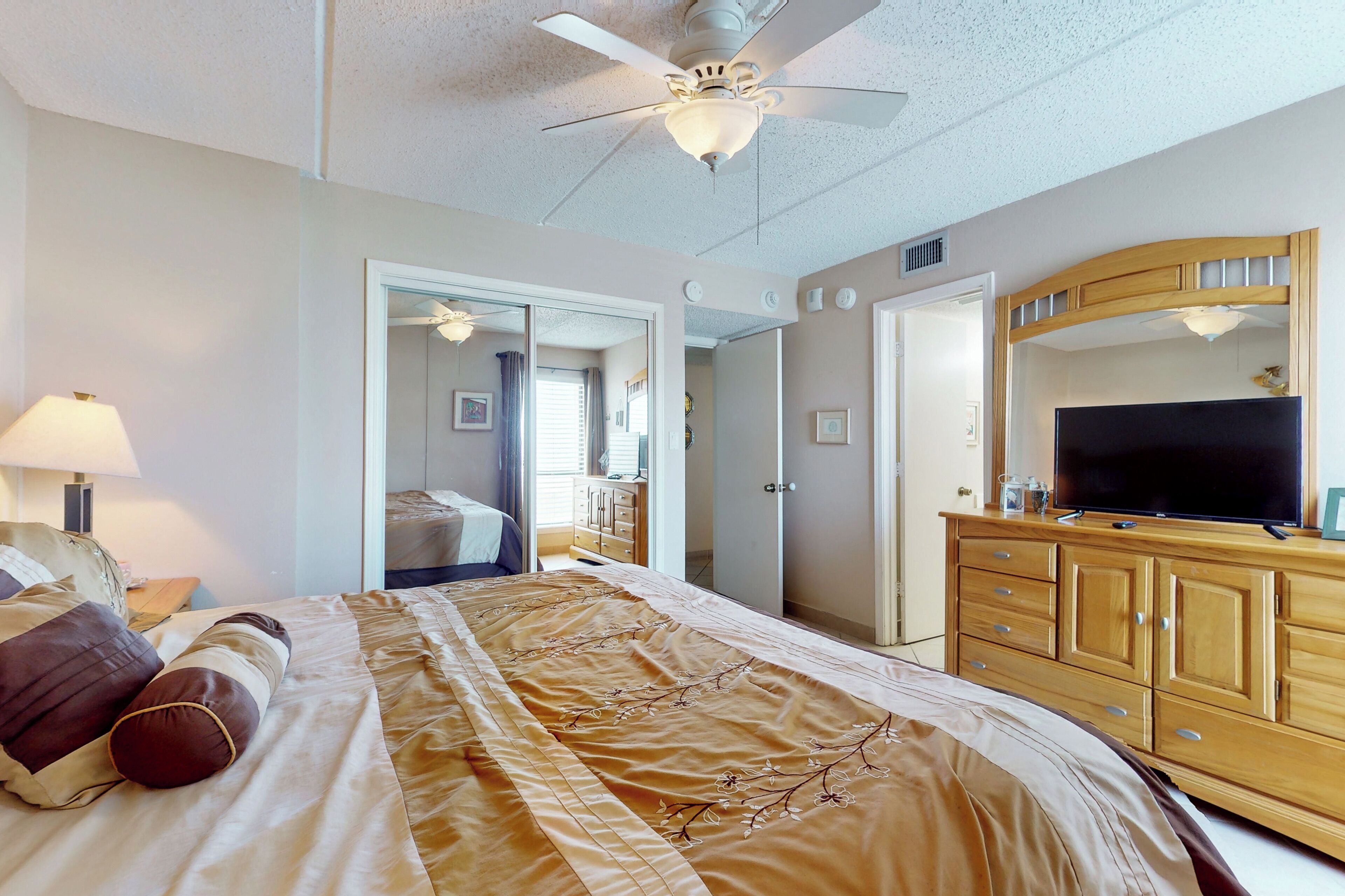 Une chambre d’hôtel avec un lit, un ventilateur de plafond, une télévision, une commode et un miroir.