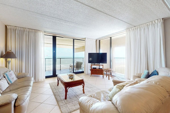Ein geräumiges Wohnzimmer mit einer großen Couch, einem Couchtisch und einem Fernseher. Ein Balkon bietet einen Ausblick.
