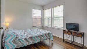 A bedroom with a bed, a TV on a wooden stand, and two windows with blinds.