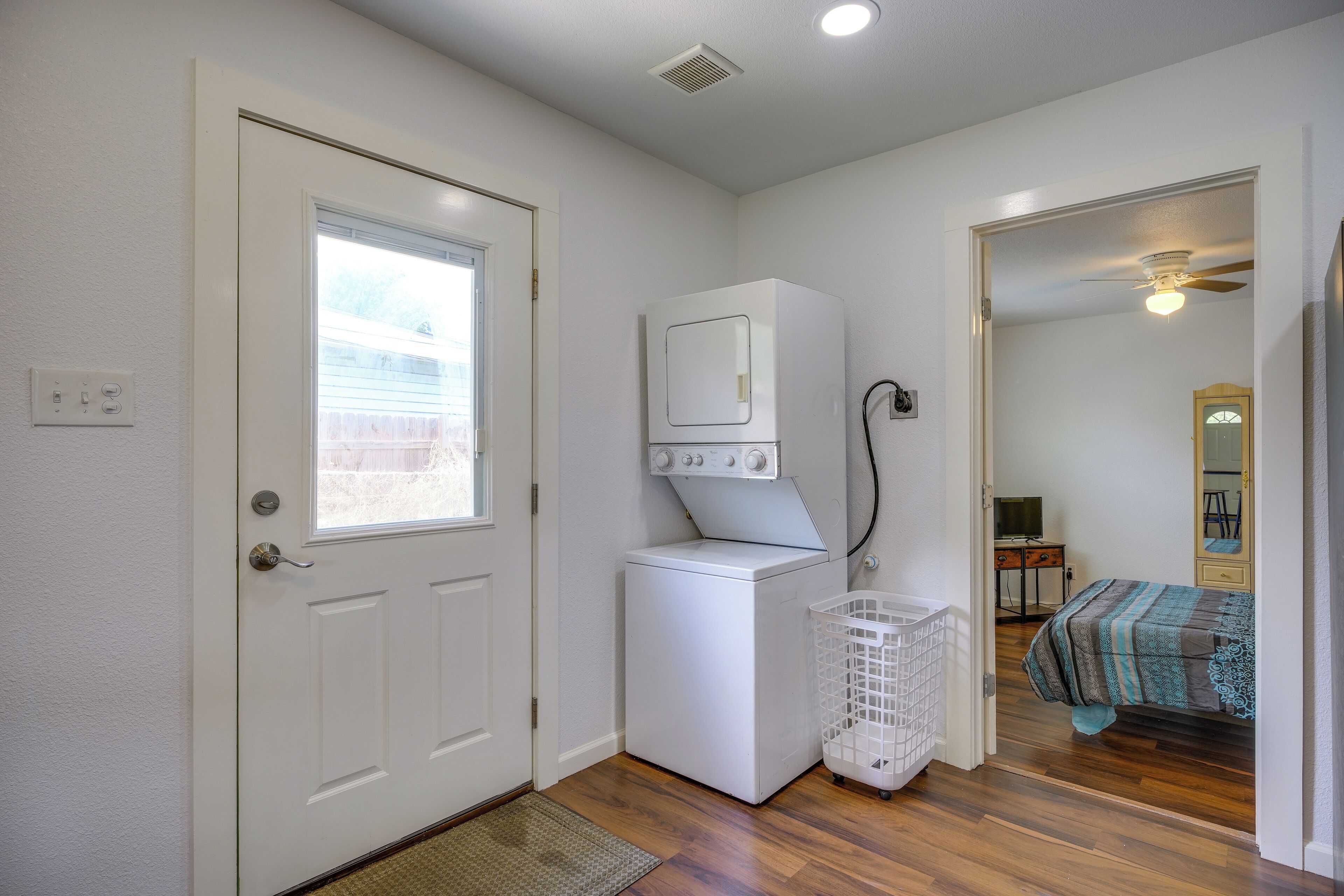 A laundry room with a washer and dryer, a basket, and a door leading to a bedroom with a bed and a ceiling fan.