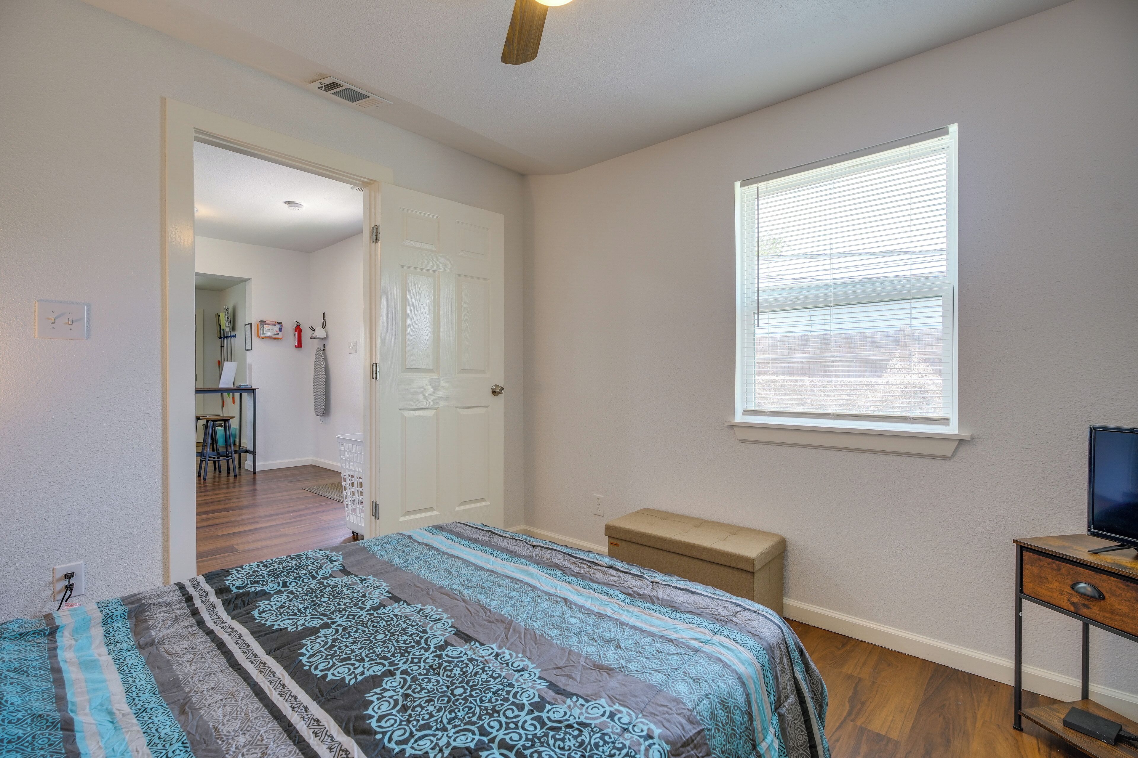 A bedroom with a bed, a desk, a window with blinds, and a door leading to another room.