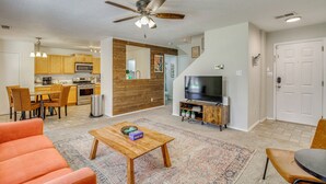 Una sala de estar con ventilador de techo, televisor, mesa de centro y comedor con sillas, y una cocina al fondo.