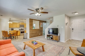 A living room with a ceiling fan, a TV, a coffee table, and a dining area with chairs and a kitchen in the background.