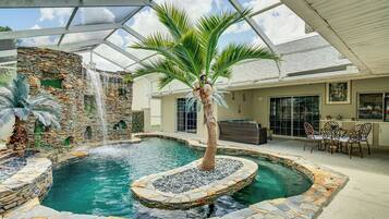 An indoor swimming pool with a waterfall, surrounded by stone walls and palm trees.
