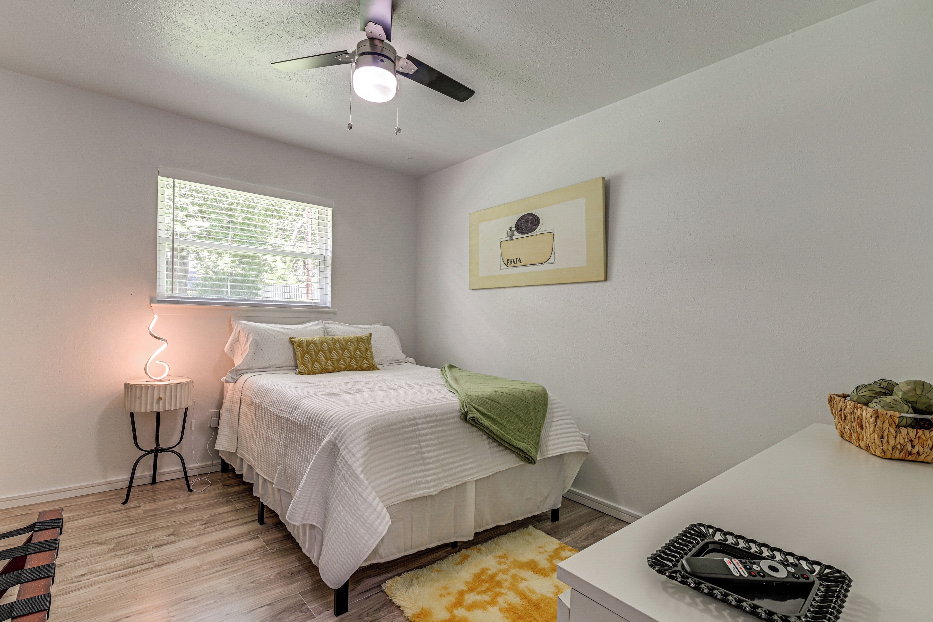 A bedroom with a bed, bedside table, lamp, ceiling fan, window, and artwork on the wall.