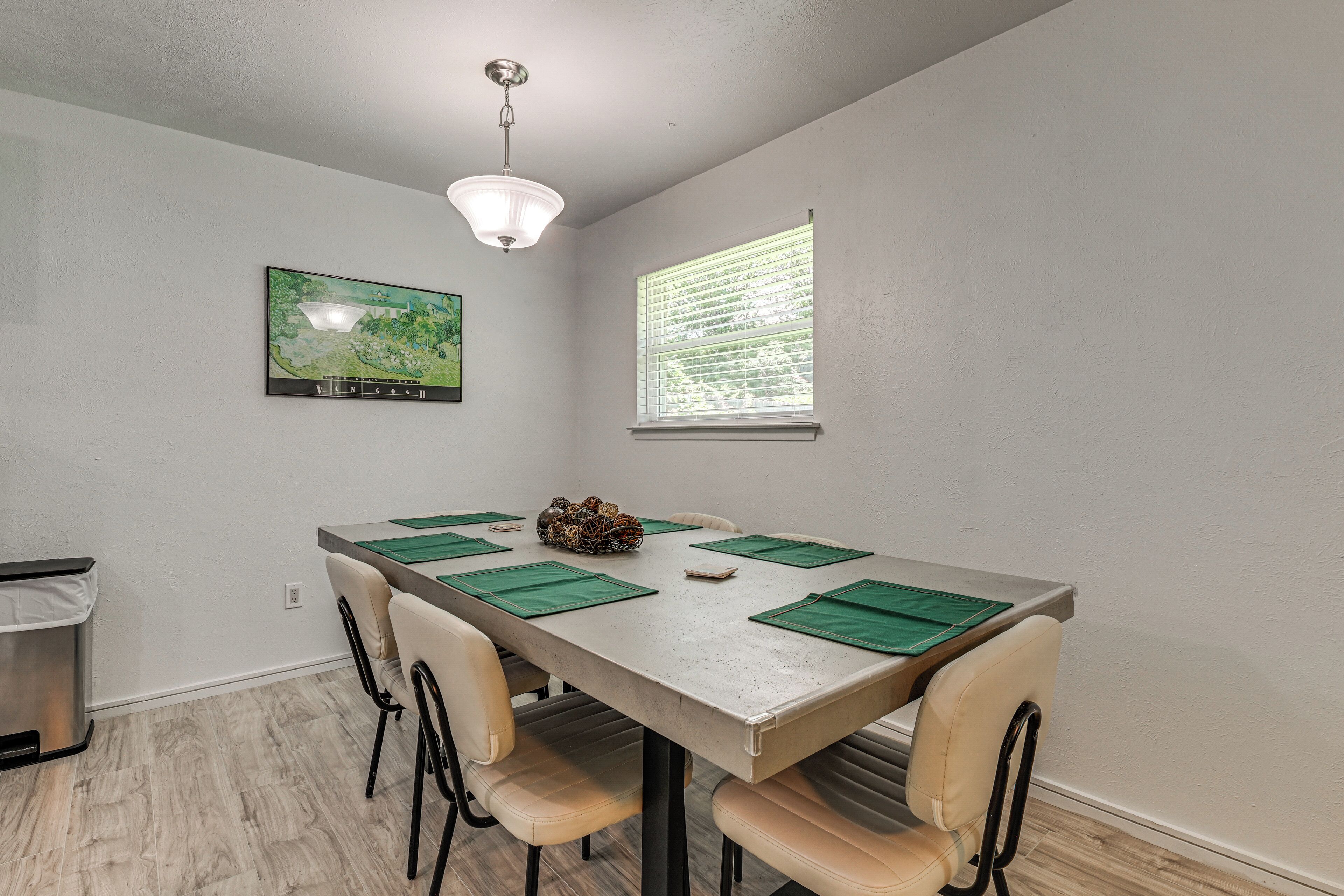 Uma sala de jantar com uma mesa, cadeiras, uma luminária no teto, uma janela e um quadro na parede.
